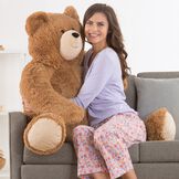 4' Big Hunka Love Bear - Seated golden brown bear with a female model in lavender pajamas on a sofa image number 0